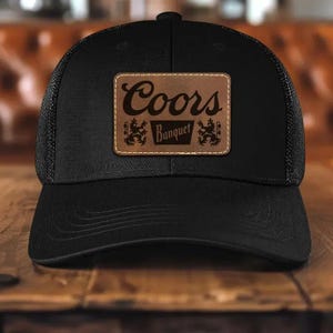 May include: Black trucker hat with a mesh back and a curved brim. The front features a rectangular brown leather patch with the words "Coors Banquet" and two decorative emblems.