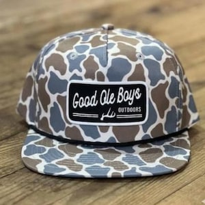 May include: A camouflage-patterned baseball cap with a flat brim. The cap features a patch with the text "Good Ole Boys Outdoors" in white on a black background. The pattern includes shades of blue, brown, and white.