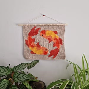 Könnte beinhalten: Ein quadratischer Wandbehang aus Häkelarbeit mit beigem Hintergrund und zwei Koi-Fischen in einem kreisförmigen Muster. Die Fische sind in Orange-, Rot- und Gelbtönen gehalten. Das Stück hängt an einem hellen Holzstab mit einer weißen Schnur.