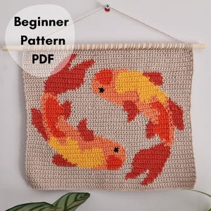 Puede incluir: Un tapiz de ganchillo cuadrado que muestra dos peces koi en formación circular. Los peces están elaborados en tonos naranja, amarillo y rojo sobre un fondo beige. Una varilla de madera sostiene la parte superior y el texto dice Beginner Pattern PDF.
