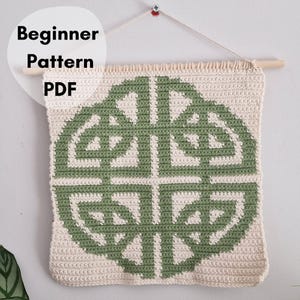 May include: A cream-colored, crocheted wall hanging with a green Celtic knot design. The text "Beginner Pattern PDF" is in a white circle in the upper left corner. The wall hanging is attached to a wooden dowel with a cream-colored string.