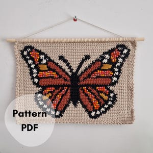 May include: A crocheted wall hanging featuring a large butterfly design. The butterfly has brown, black, orange, and white details. The text "Pattern PDF" is visible on the lower left. The wall hanging is attached to a wooden dowel.