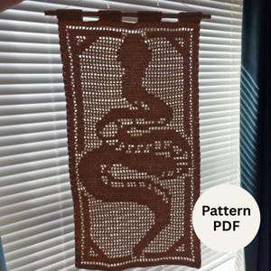 May include: A rectangular brown crochet wall hanging featuring a detailed snake silhouette design. The piece is suspended from a thin wooden rod by four crochet loops at the top. The intricate openwork pattern creates a clear image of a coiled snake.