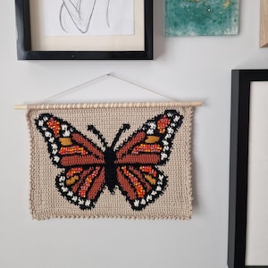 May include: A rectangular crochet wall hanging featuring a monarch butterfly design in shades of orange, black, and white on a beige background. The textile is suspended from a light-colored wooden dowel with a thin white string for hanging.