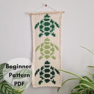 Op de afbeelding: Een crèmekleurige, rechthoekige gehaakte wandhanger met drie groene schildpadontwerpen. De wandhanger is opgehangen aan een houten roede. De tekst "Beginner Pattern PDF" is zichtbaar in de linker benedenhoek.