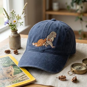 Peut inclure: Casquette de baseball bleu marine avec un motif brodé de deux singes. L'un est allongé, l'autre debout et tenant un objet. La casquette est en tissu doux et texturé.