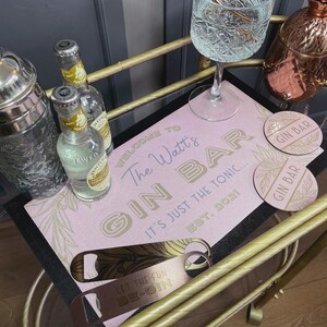 Puede incluir: Una barra de gin rosa con una alfombrilla decorativa que dice "Welcome to The Watt's Gin Bar". Incluye dos botellas de agua tónica, dos posavasos y dos abridores de botellas. El texto "Let the fun begin" está en un abridor.