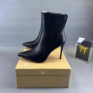 May include: A pair of black leather ankle boots with a pointed toe and a high stiletto heel. The boots feature a side elastic panel and are displayed on a tan cardboard box with the word Paris printed on the side and a script logo on the front.