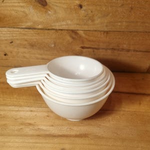 May include: A stack of white measuring cups, made of plastic. The cups are nested together, with handles for easy use. The set includes several different sizes for accurate measurements in the kitchen. The cups are in a light colour.