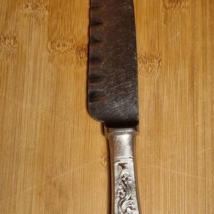 May include: A stainless steel serrated knife with a decorative handle. The blade has a curved shape. The handle is silver-coloured and features an embossed floral design. The knife is on a wooden chopping board.