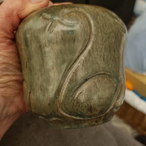 May include: A small, rounded ceramic vessel with a matte, mottled green glaze. The surface features a raised, stylized swan design. The vessel is held in a hand, suggesting its size and form.