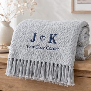 Può includere: Una coperta grigio chiaro con un motivo a spina di pesce testurizzato e lunghe frange. La coperta presenta un testo ricamato in blu scuro che recita J cuore K e la frase Our Cozy Corner centrata sulla superficie anteriore del tessuto.