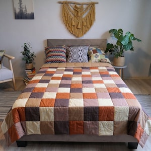 Könnte beinhalten: Eine Patchwork-Tagesdecke mit einem Raster aus quadratischen Stoffbahnen in abwechselnden Farbtönen von gebranntem Orange, Creme, Dunkelbraun und gedecktem Mauve. Die Decke liegt auf einem Bett mit grauem Polsterkopfteil und drei Zierkissen.