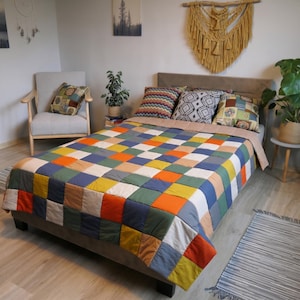 Op de afbeelding: Een patchwork sprei met een raster van vierkante stoffen lappen in diverse kleuren, waaronder oranje, blauw, groen, geel en beige. De sprei ligt op een bed met een beige hoofdeinde, vergezeld door drie sierkussens met geometrische patronen.