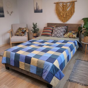 May include: A patchwork quilt featuring a grid of square fabric patches in various shades of blue, light blue, and beige. The quilt is draped over a bed with a brown headboard, accompanied by several decorative pillows with geometric patterns.