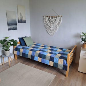 May include: A cozy bedroom scene featuring a wooden bed frame with a patchwork quilt in shades of blue, green, and tan. Two pillows rest on the bed. A macrame wall hanging and two landscape paintings add to the room's decor.