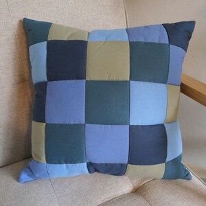 Könnte beinhalten: Ein quadratisches Zierkissen mit einem Patchwork-Design, das aus mehreren Stoffquadraten in verschiedenen Blau-, Marine- und Olivgrüntönen besteht. Das Kissen ist mit einem Gittermuster aus vernähten Quadraten konstruiert, was eine gleichmäßige Oberfläche ergibt.