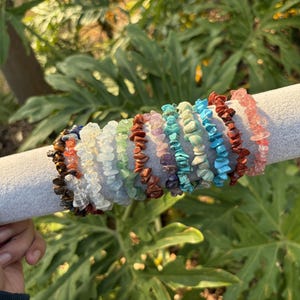 May include: A collection of gemstone chip bracelets in various colors, including red, blue, green, and pink. The bracelets are displayed on a gray cylindrical holder against a backdrop of green foliage.