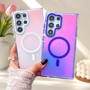 Op de afbeelding: Twee transparante telefoonhoesjes met een roze en paarse gradiënt. Elke hoes heeft een cirkelvormig wit of paars accent en een camera-uitsparing. De hoesjes worden voor een vage achtergrond gehouden.