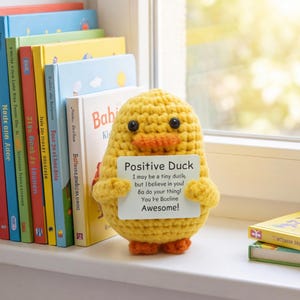May include: A yellow crocheted duck plush toy with black bead eyes and an orange beak. The duck holds a white rectangular sign that reads: Positive Duck, I may be a tiny duck, but I believe in you! Go do your thing! You're Awesome!