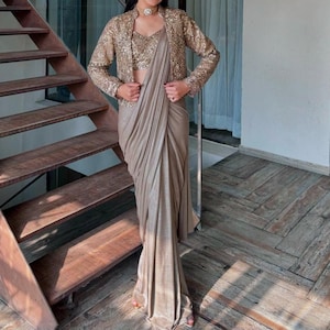 Op de afbeelding: Een metallic champagnekleurige sari gecombineerd met een bijpassende mouwloze top en een jasje met lange mouwen dat zwaar is bezet met lovertjes. Het ensemble heeft een gedrapeerde rok met een geplooid voorpand en een glanzende stofafwerking.