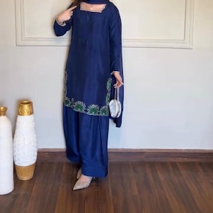 May include: A deep navy blue long-sleeved tunic and matching loose-fitting trousers. The tunic features a square neckline and a hemline decorated with green and white floral embroidery. A matching navy blue scarf and a small silver handbag are included.