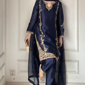 May include: A dark navy blue satin salwar kameez featuring intricate gold embroidery along the neckline, side panels, and sleeve cuffs. The ensemble includes a matching sheer navy blue dupatta with subtle embellishments and coordinating trousers.