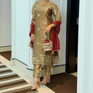 May include: A metallic gold long-sleeved tunic and matching trousers set featuring intricate gold embroidery and sequin embellishments. A deep red sheer shawl with gold borders is draped over the arms, paired with a matching red and gold handbag.