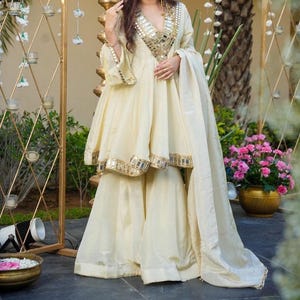May include: A cream-coloured three-piece outfit consisting of a flared tunic with a deep V-neckline, wide-leg trousers, and a matching dupatta. The tunic features intricate mirror work embroidery along the neckline, cuffs, and hemline.