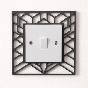 May include: A white light switch with a rectangular, gray geometric frame. The frame has a repeating pattern of triangles and lines. The switch plate is secured with two small screws.