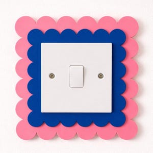 以下が含まれることがあります： 長方形のトグルが付いた白いスイッチ。装飾フレーム内に設定されています。フレームは、ピンクと青の層を備えたスカラップエッジデザインが特徴です。スイッチは白い壁に取り付けられています。