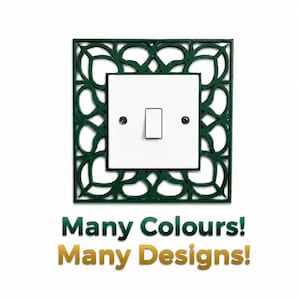Op de afbeelding: Een vierkante witte lichtschakelaar met een donkergroen decoratief frame met een ingewikkeld uitgesneden patroon. De tekst onder de schakelaar luidt Many Colours! Many Designs! De schakelaar is bevestigd met twee zichtbare zilveren schroeven.