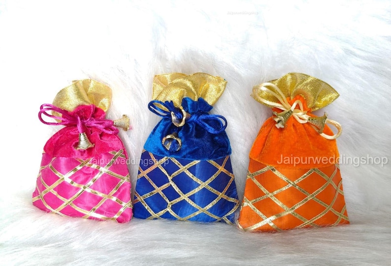 May include: Three colorful velvet drawstring bags. The bags are pink, blue, and orange, with gold accents. Each bag has a gold top and a gold lattice pattern on the bottom. The bags are tied with a matching drawstring.