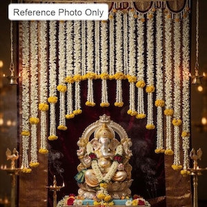 Puede incluir: Una estatua metálica de Ganesha sobre un pedestal frente a un fondo de tela roja oscura adornado con guirnaldas de flores blancas y racimos de caléndulas amarillas. Dos lámparas de aceite de latón, con el texto Reference Photo Only.