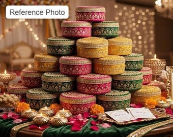 May include: A pyramid stack of round, lidded boxes in vibrant shades of pink, green, and yellow, each featuring intricate gold embroidery and decorative trim. The boxes are arranged on a table with scattered flower petals and small tea light candles.