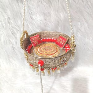 Op de afbeelding: Een ronde, goudkleurige decoratieve mand met ingewikkeld metalen gaas, hangend aan twee met parels versierde kettingen. De binnenkant bevat twee rode vierkante kussens met gouden borduursels en een centraal gouden rond medaillon met rode pompons.
