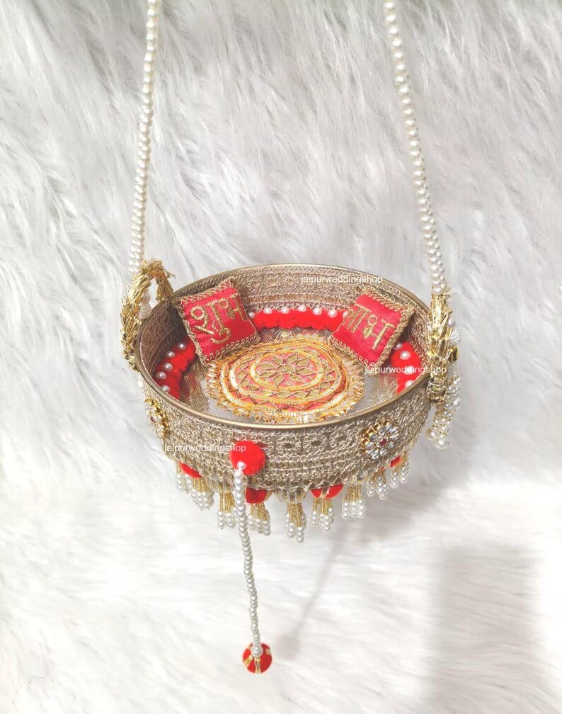 Op de afbeelding: Een ronde, goudkleurige decoratieve mand met ingewikkeld metalen gaas, hangend aan twee met parels versierde kettingen. De binnenkant bevat twee rode vierkante kussens met gouden borduursels en een centraal gouden rond medaillon met rode pompons.