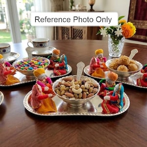 May include: A table setting featuring silver serving trays with bowls of colorful candies, nuts, and sweets. Miniature figures in traditional attire surround each bowl. A vase of flowers and teacups are also present.