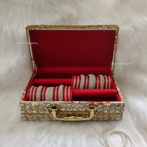 Puede incluir: Una caja de joyería abierta y ornamentada con un interior de terciopelo rojo, que contiene múltiples juegos de brazaletes plateados y rojos. La caja está decorada con bordados florales dorados y blancos y tiene un asa dorada. El texto "Jaipurweddingshop" es visible.
