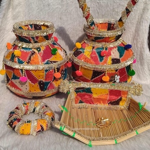 Puede incluir: Un conjunto de artículos de boda indios hechos a mano. El conjunto incluye dos ollas decoradas, una bandeja pequeña y un objeto circular, todos adornados con patchwork vibrante, adornos dorados y pompones. Estos artículos se utilizan en ceremonias tradicionales.