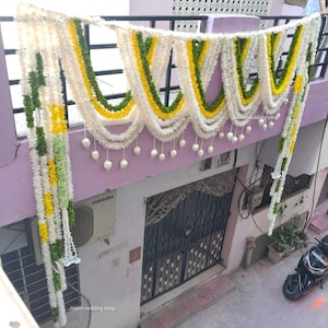 May include: A decorative garland of white, yellow, and green faux flowers hangs above a doorway. The garland is draped in a swag pattern and decorated with white teardrop-shaped ornaments. The scene is set outdoors, with a glimpse of a building and a motorcycle.