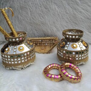 May include: Two silver-toned metal pots decorated with gold trim, small circular mirrors, and white beaded fringe. A matching gold-trimmed triangular tray sits between them, accompanied by two circular bangles wrapped in pink and gold fabric.