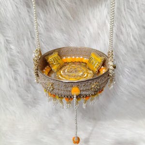 Op de afbeelding: Een ronde goudkleurige decoratieve schommel met ingewikkelde filigraanpatronen, versierd met gele stoffen kussentjes, kleine gele pompons en witte parelachtige strengen die aan de rand hangen, opgehangen aan twee lange parelkettingen.