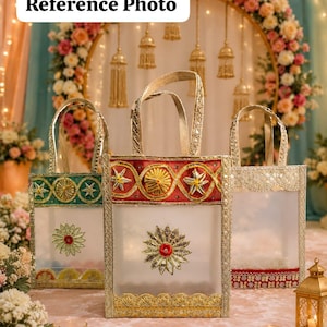 Può includere: Tre borse rettangolari con pannelli trasparenti e manici dorati. Le borse presentano bordi in tessuto decorativo in verde, rosso e oro con motivi floreali ricamati, paillettes e finiture metalliche. Ogni borsa ha un design strutturato.
