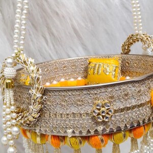 Op de afbeelding: Een ronde decoratieve mand met een metallic zilveren band met ingewikkeld goudborduurwerk en een centraal bloemmotief. De rand is versierd met gele en oranje stoffen pompons, parelkwastjes en twee met parels bezette handvatten.