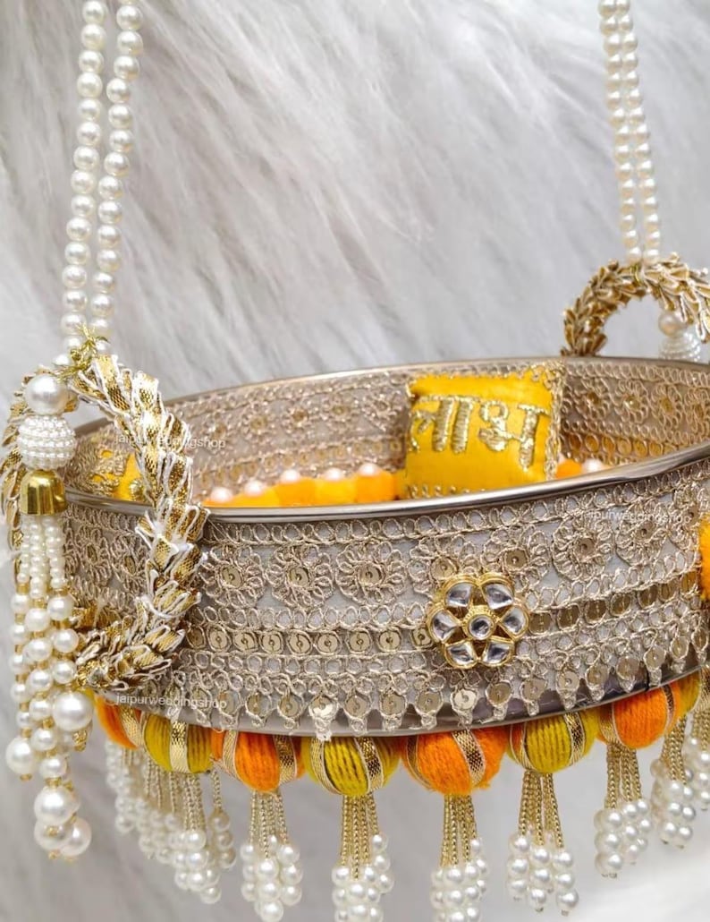 Op de afbeelding: Een ronde decoratieve mand met een metallic zilveren band met ingewikkeld goudborduurwerk en een centraal bloemmotief. De rand is versierd met gele en oranje stoffen pompons, parelkwastjes en twee met parels bezette handvatten.