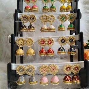 May include: A display of colorful, ornate earrings. Each pair features a floral design with a central gem and a dangling bell shape. The earrings come in various colors, including red, green, yellow, and pink, with gold-colored accents and small pearl-like beads.