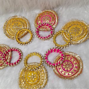 Op de afbeelding: Een set van vijf ronde stoffen onderzetters en vijf bijpassende armbanden. De onderzetters en armbanden zijn gemaakt van felgele of felroze stof met goudkleurig borduurwerk, spiegelachtige accenten en een rand van kleine witte kraaltjes.