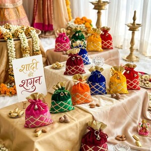 Può includere: Una collezione di sacchetti regalo in velluto colorato con dettagli dorati e motivi a rombi. I sacchetti sono disposti su un tavolo con mandorle e piccole statuette decorative. L'immagine include un cartello con testo in lingua straniera.