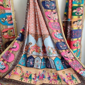 May include: A colorful textile with intricate patterns and designs. The fabric features a mix of pink, blue, orange, and gold hues, with detailed illustrations of figures and scenes. The edges are trimmed with gold accents and the text "Jaipurweddingshop" is visible.